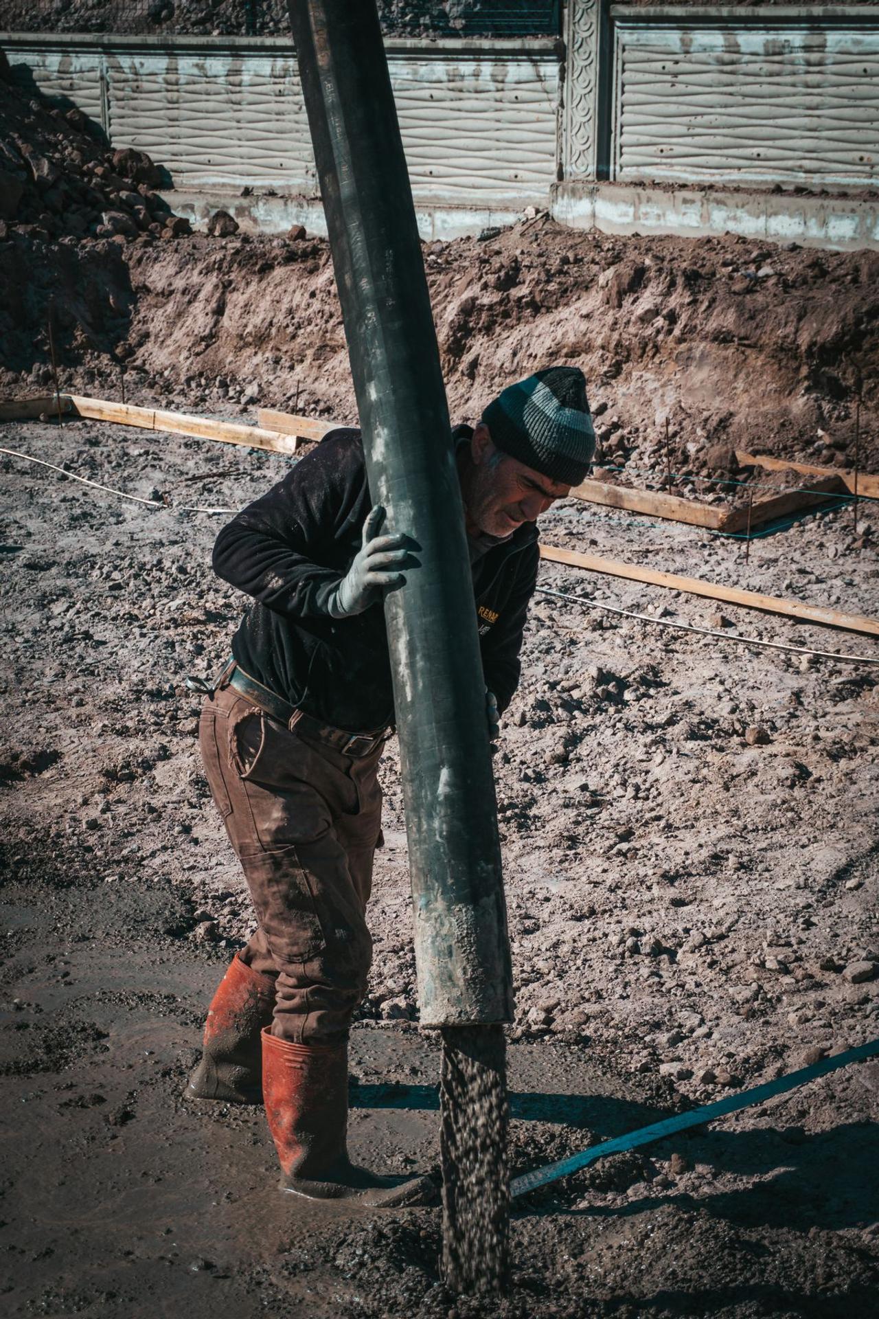Worker in a beanie guiding a concrete pump hose while pouring a footing trench at a job site