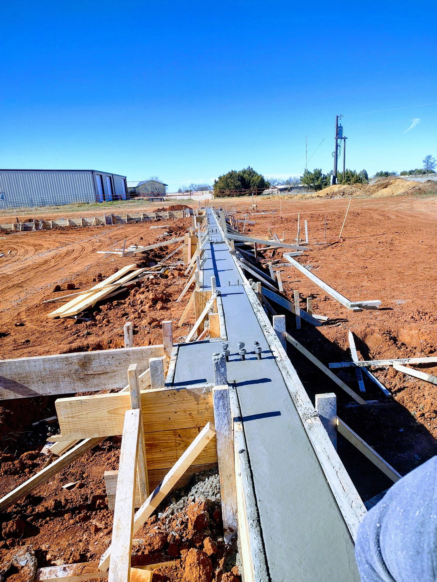 Long concrete foundation footer freshly poured within wood formwork, stretching toward a metal building on a clear winter day