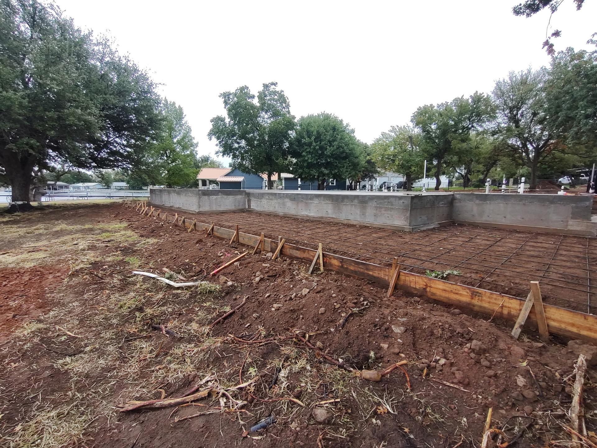 Poured concrete foundation wall and rebar slab grid on a lakeside lot, with homes visible across the water