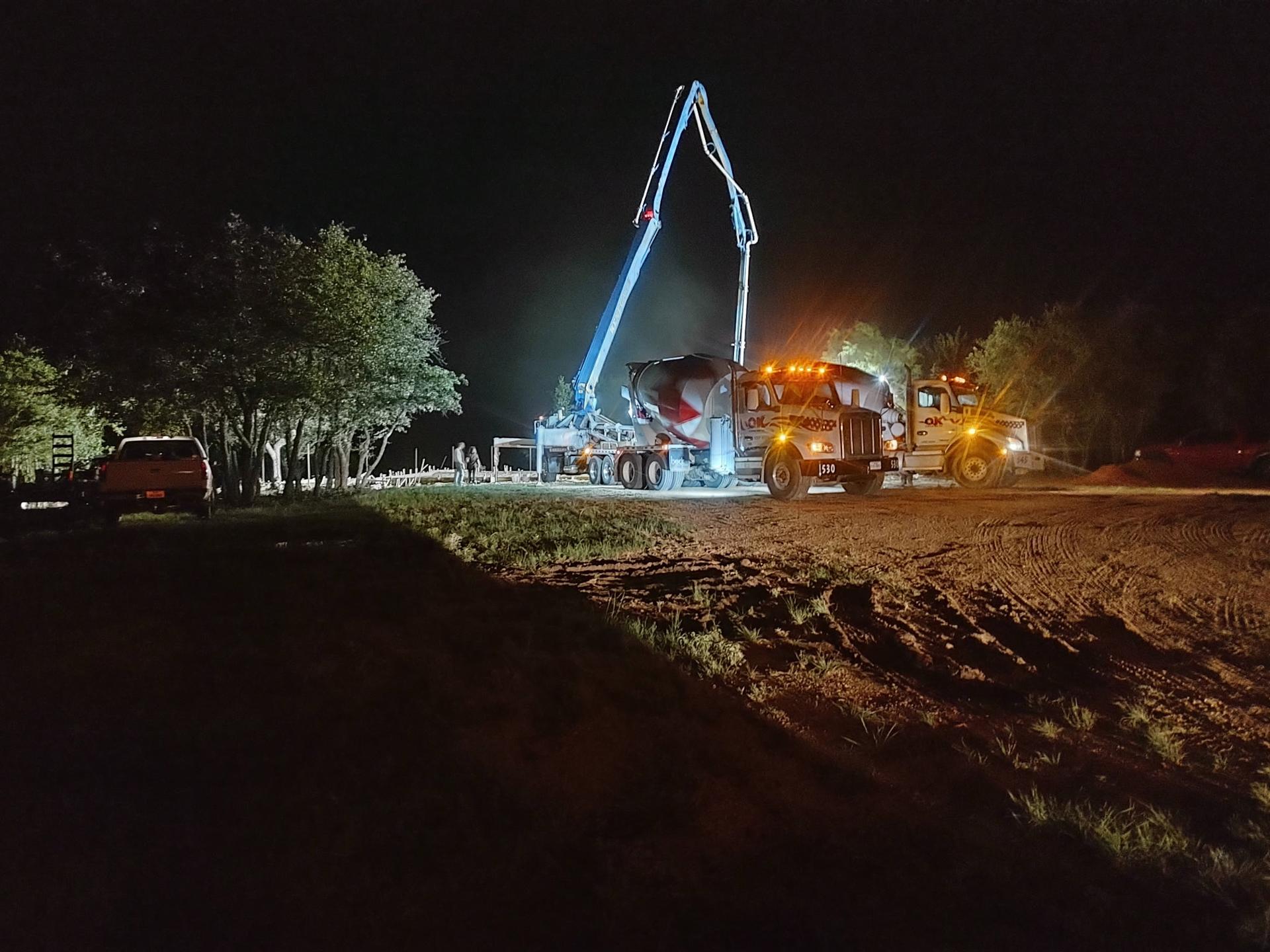 Two concrete mixer trucks parked with the pump boom extended skyward at a rural jobsite at night