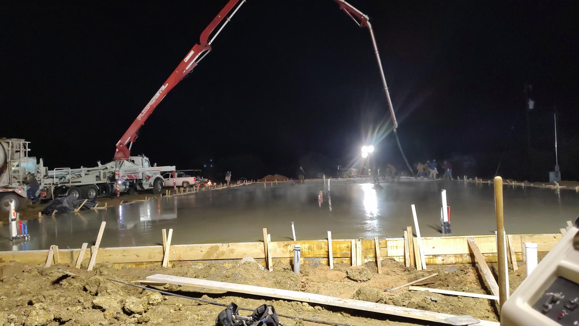 Crew and pump truck pouring a concrete slab at night under floodlights, with the mixer truck staged alongside