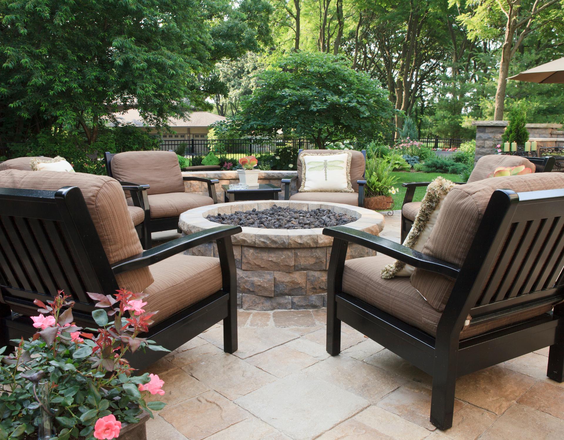 Paver patio with a built-in stone fire pit surrounded by cushioned outdoor chairs in a landscaped backyard.