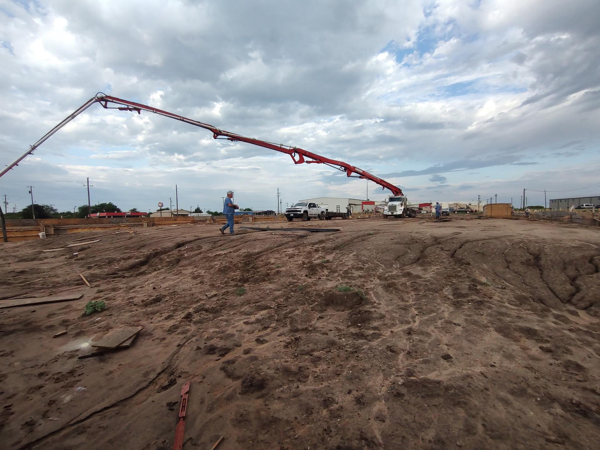 Concrete pump truck boom extended over a graded, muddy commercial building site under an overcast sky