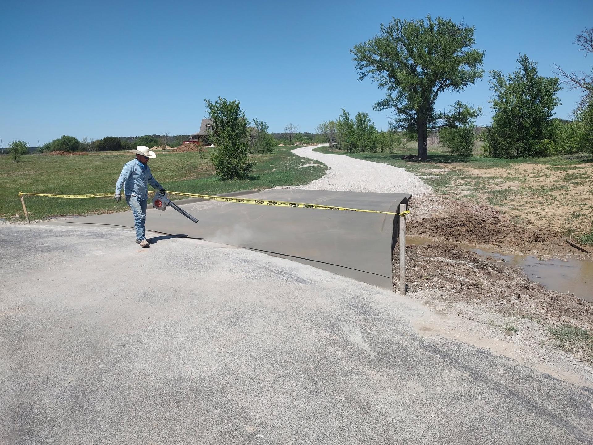 Concrete worker in a cowboy hat clearing dust off a freshly poured rural driveway crossing