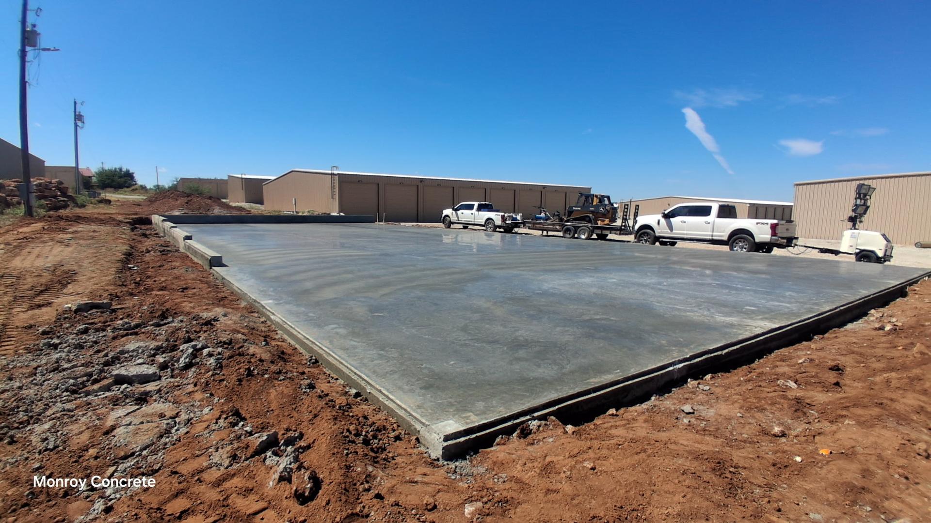 Freshly poured concrete slab in front of a row of metal shop buildings, with pickup trucks and a skid steer staged nearby