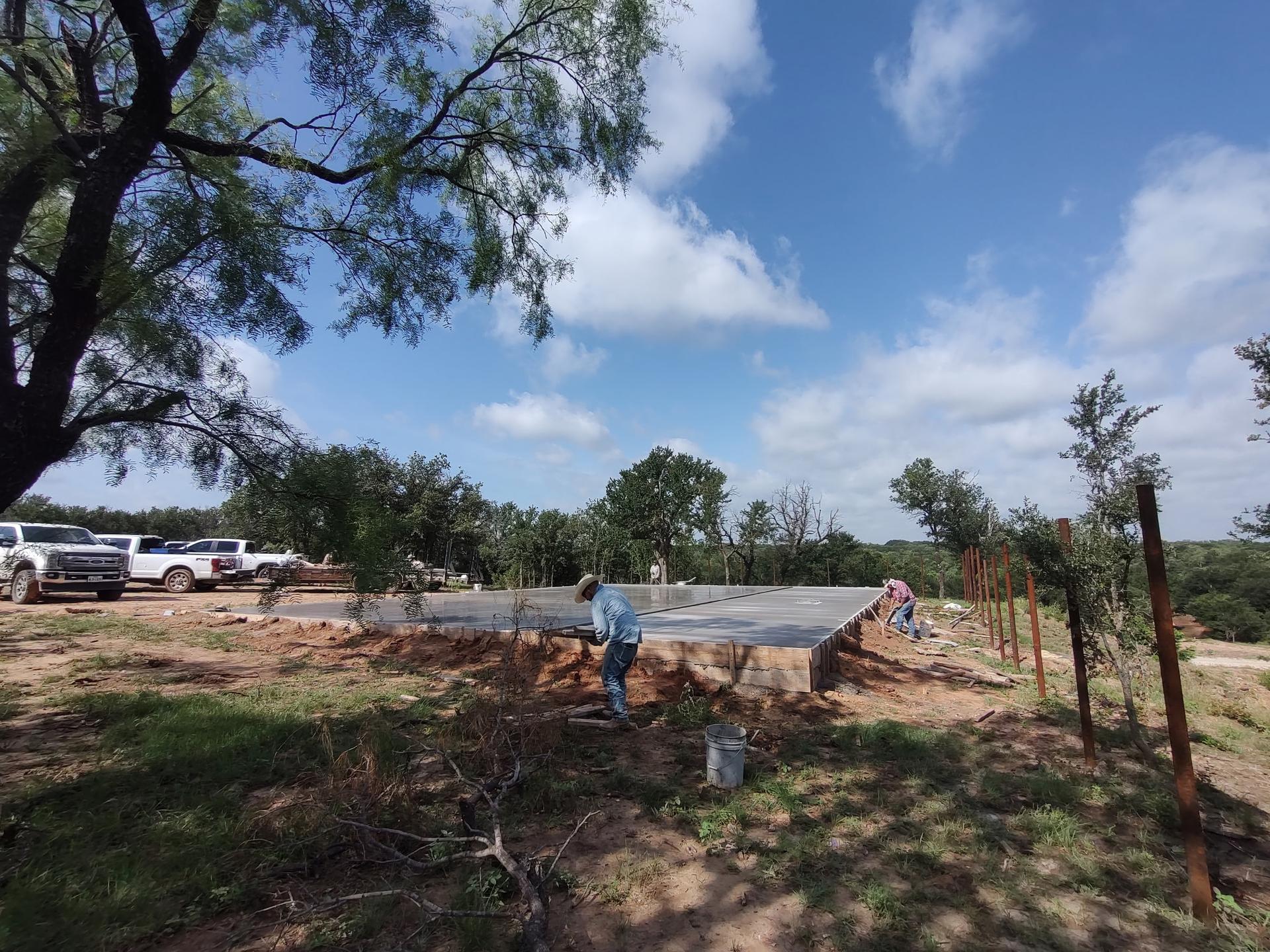 Two workers hand-finishing the edge of a concrete slab on a wooded rural lot, with rebar posts staged for a fence line nearby
