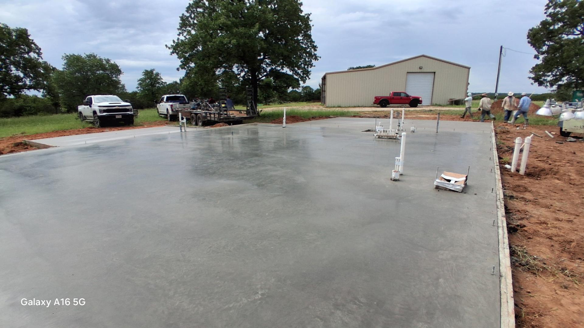 Freshly finished concrete slab with plumbing stub-outs in front of a metal building, crew and pickup trucks visible in the background
