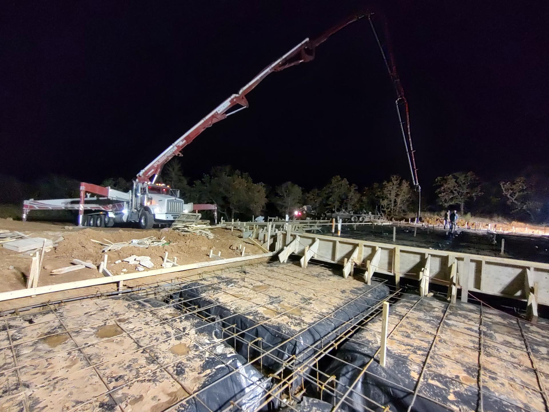 Crew pouring and finishing a concrete slab under work lights at night, with the pump truck boom extended overhead