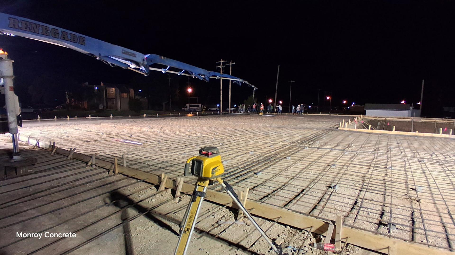 Laser level set up on a large rebar-reinforced concrete slab poured at night, with the pump truck boom extended and crew working in the background