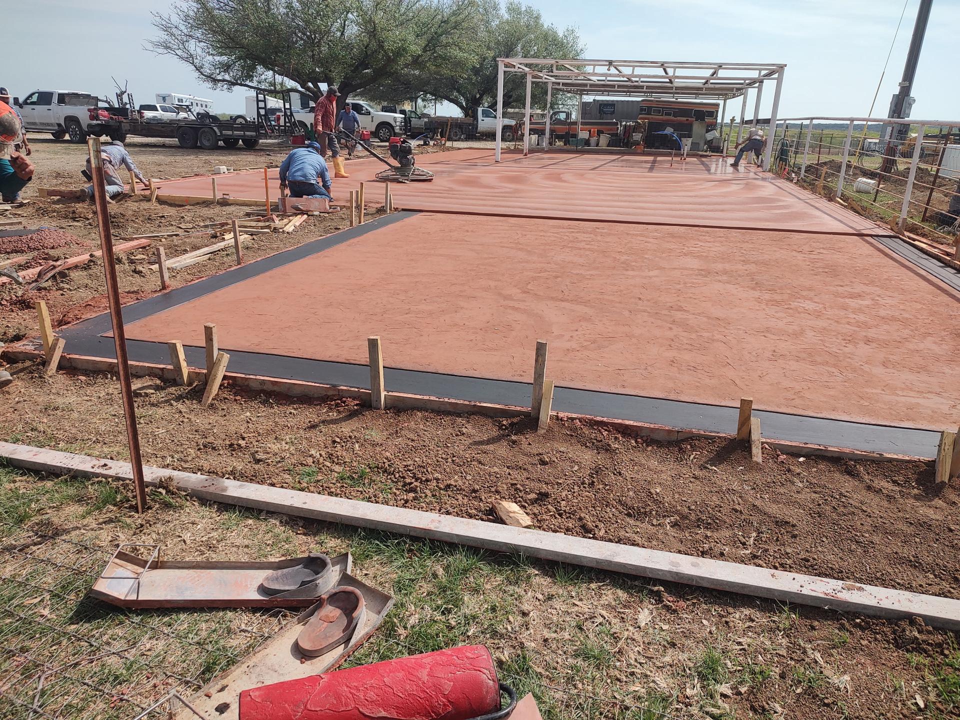 Freshly finished red-tinted concrete patio slab beneath a white pergola frame on a ranch property, with crew power troweling the surface